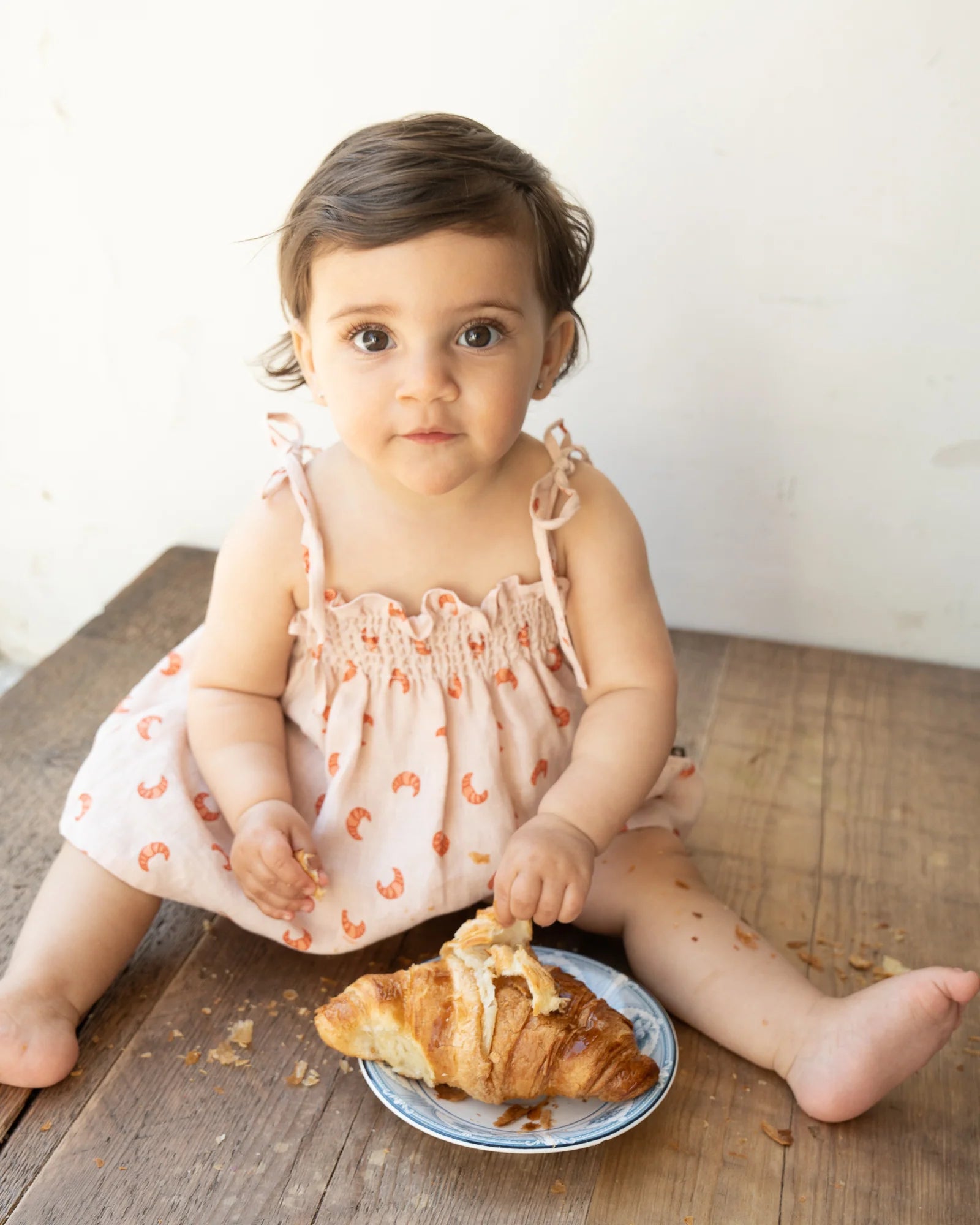 A toddler wearing a light pink dress sits on a wooden surface, holding and eating a croissant from a small blue and white plate. Croissant crumbs are scattered around.