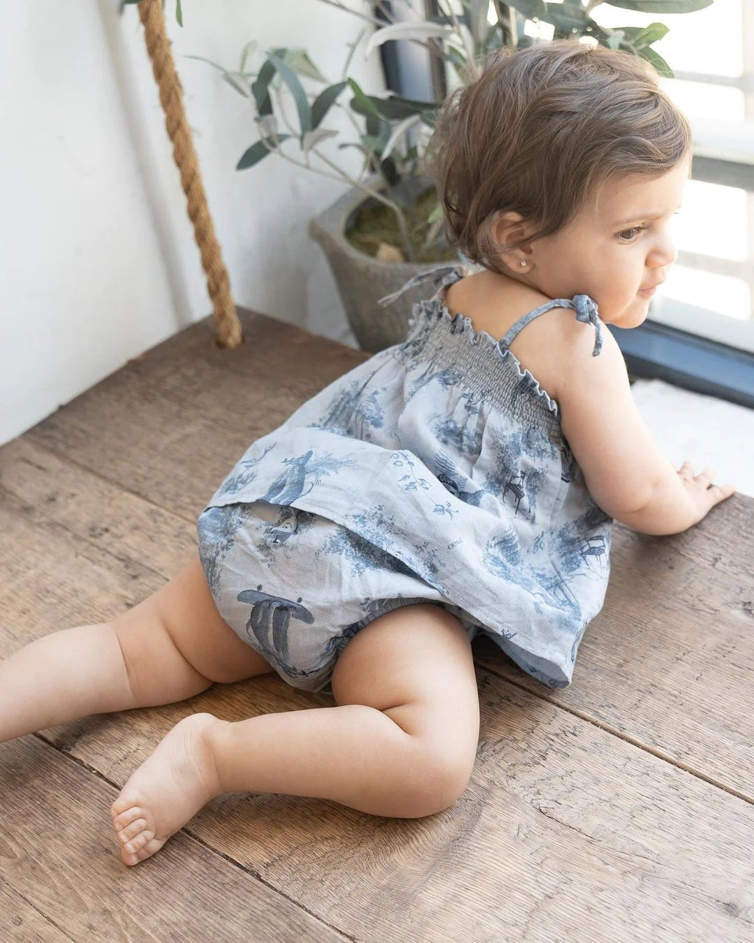 A baby wearing a blue patterned dress and diaper is crawling on a wooden floor near a window, with a potted plant in the background.