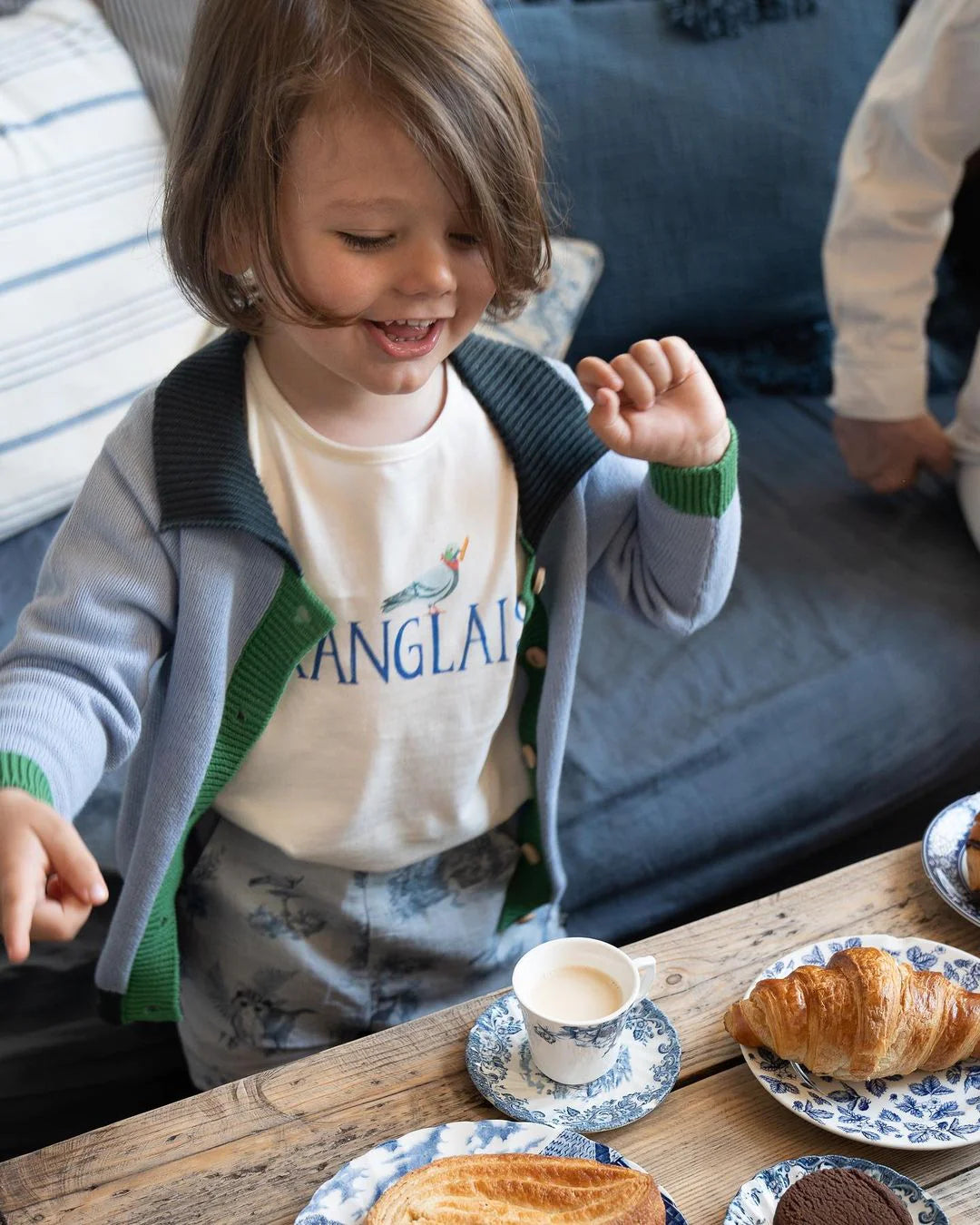 Child with shoulder-length hair, wearing a cardigan and printed shirt, excitedly stands by a table with pastries and a cup on blue-patterned plates.