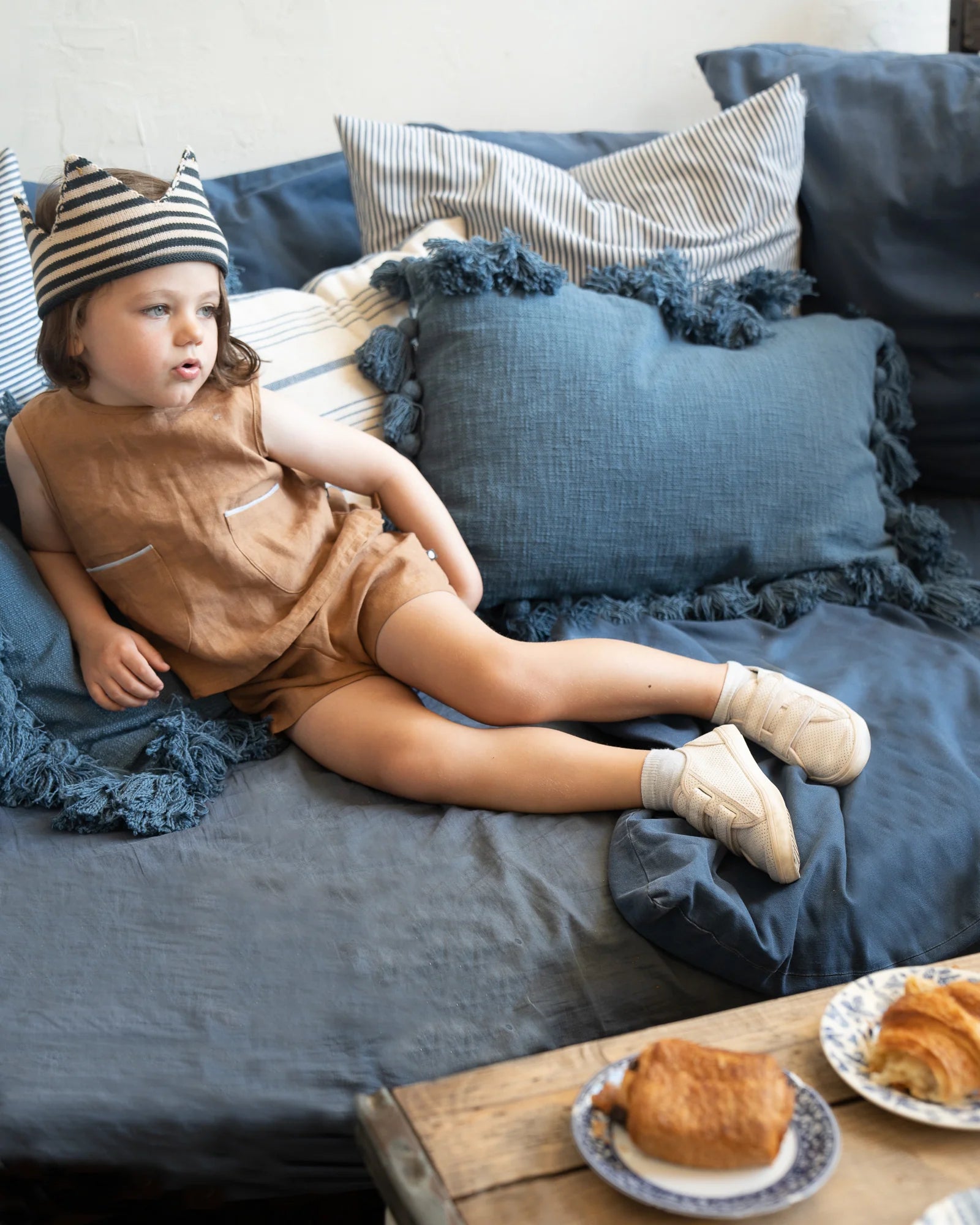 Child in brown outfit and striped hat lounges on a blue couch, with croissants on a wooden table in the foreground.
