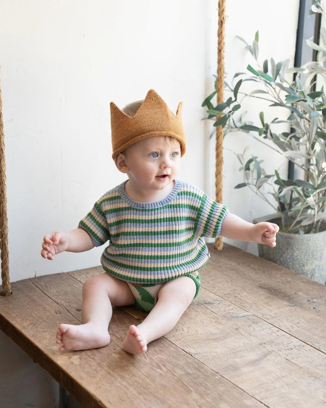 A baby wearing a knitted crown and striped outfit sits on a wooden surface near a plant, looking sideways.