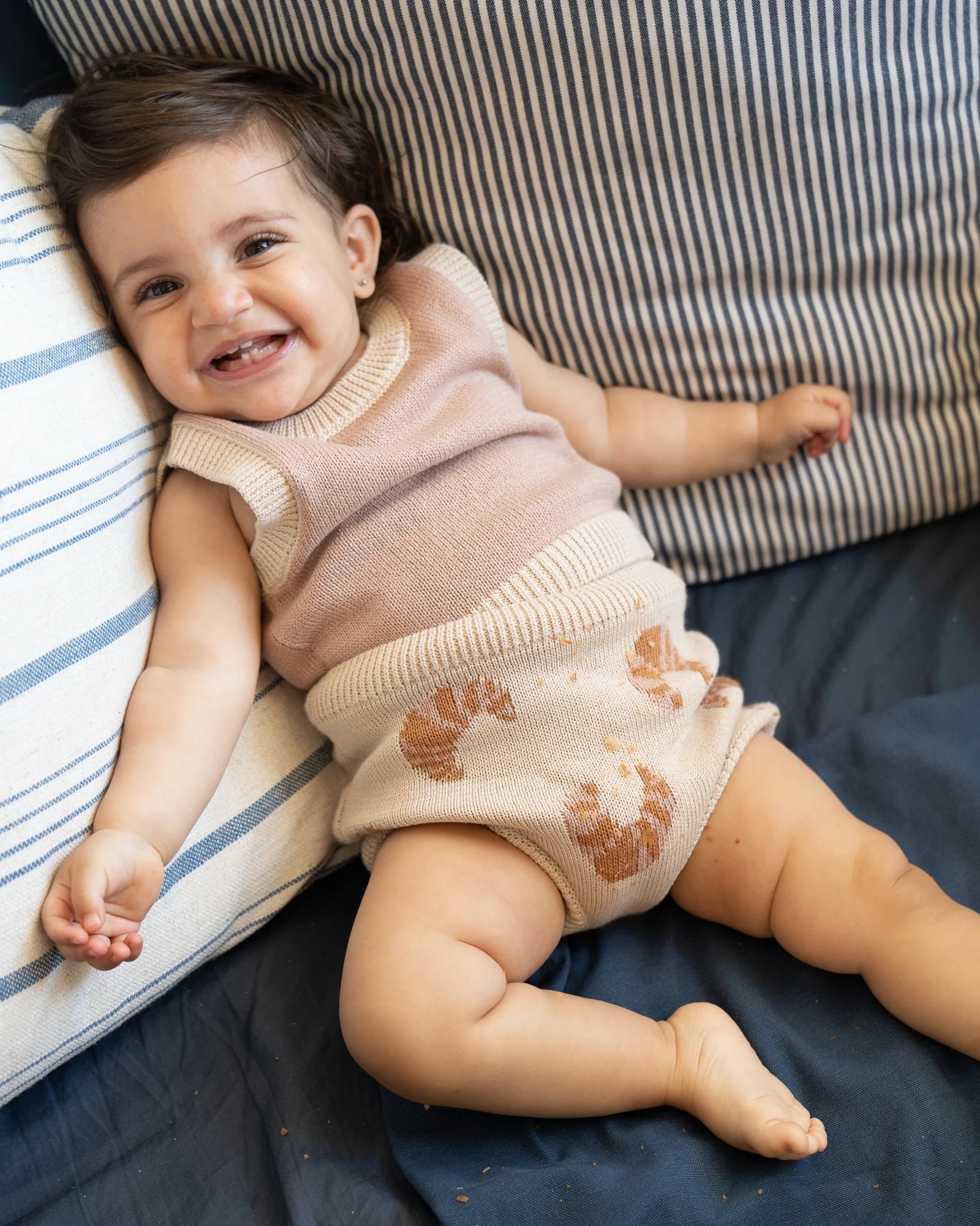 Smiling baby lying on a navy blue surface wearing a light brown sleeveless top and matching shorts with design, surrounded by striped pillows.