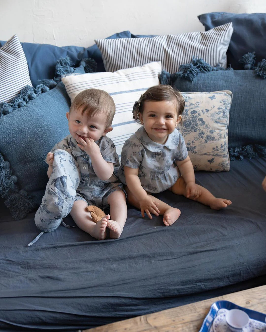 Two toddlers in matching outfits sit on a blue couch with patterned cushions, smiling and looking at the camera.