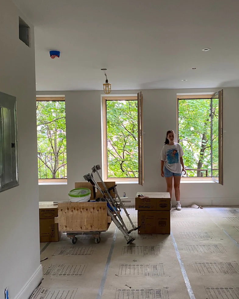 Person standing in a partially furnished room with open windows and boxes. Trees are visible outside.
