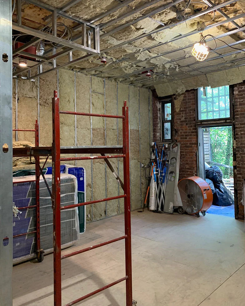 Construction site with exposed brick walls, metal studs, and insulation. A red scaffold is in the center, with tools and materials like ladders and a large fan in the background.