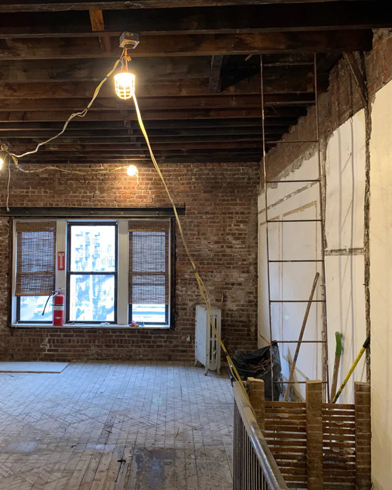 Industrial-style room under renovation with exposed brick walls, wooden beams, hanging lights, and a large window. Building materials and tools are scattered on the floor.