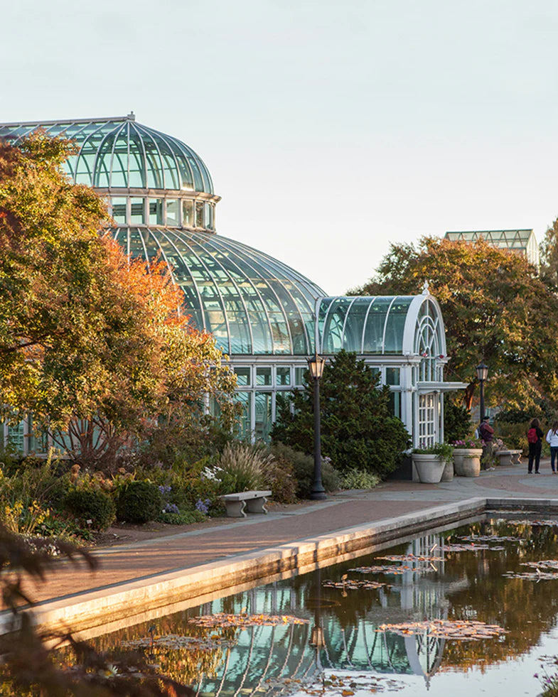 A glass-domed conservatory with surrounding trees and a pond reflecting the structure. People walk along the pathway.