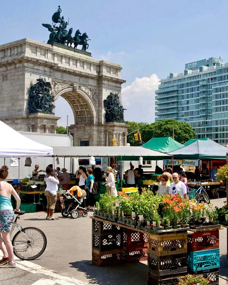 People browse a farmers market near a large stone arch with a sculpture on top, surrounded by tents and stalls with plants and produce, on a sunny day in an urban setting.