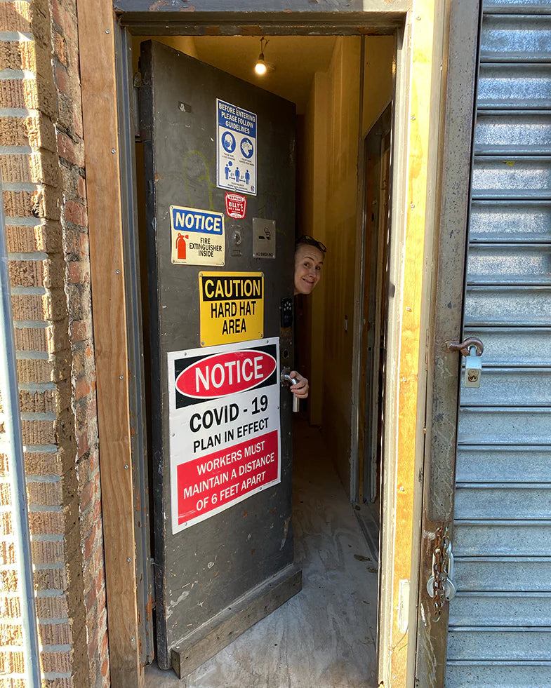 A person peeks from a partially open door covered with various warning notices including COVID-19 precautions and hard hat area signs.