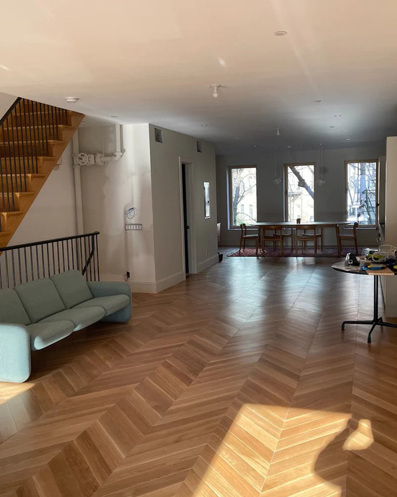 Spacious living area with wooden chevron-pattern floor, light green sofa, wooden staircase, table with chairs, and large windows.