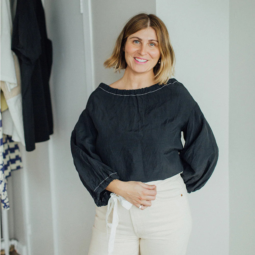 A person with shoulder-length hair smiles, wearing a black blouse and light-colored pants, standing indoors.