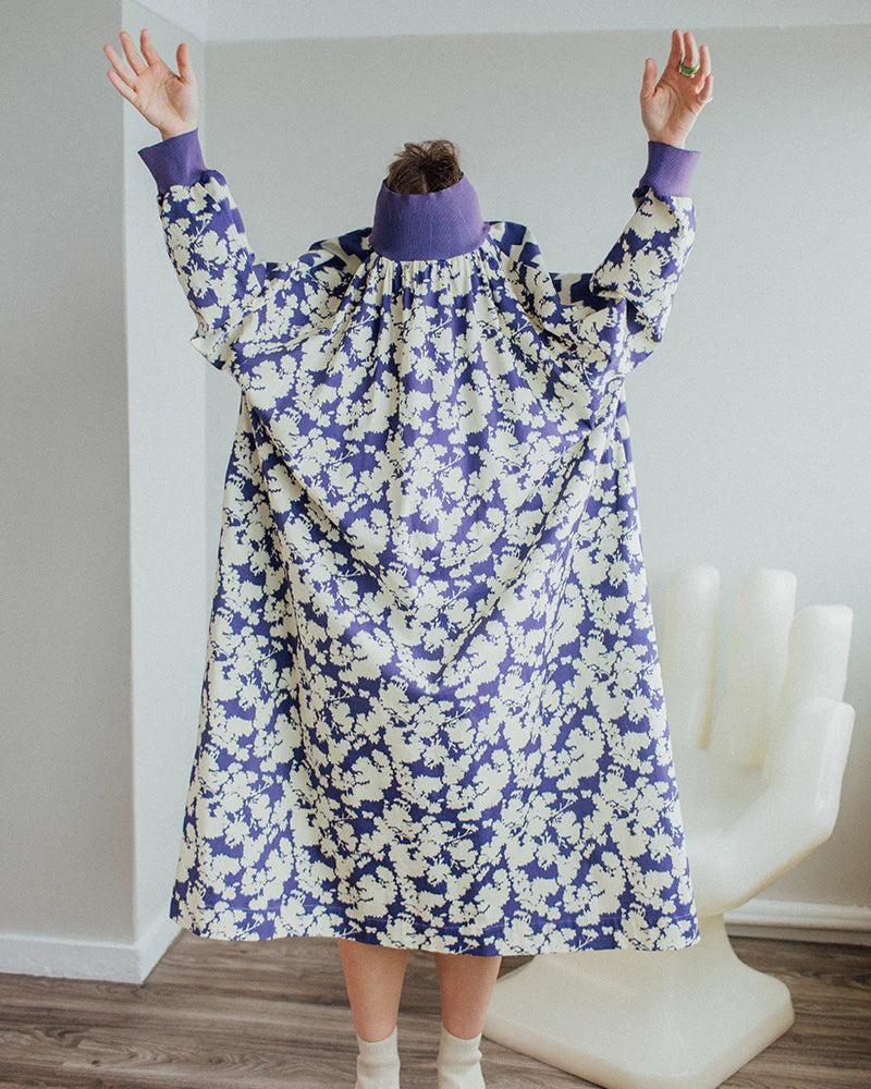 Person with head inside purple and white floral garment, raising arms. Background includes a white chair shaped like a hand.