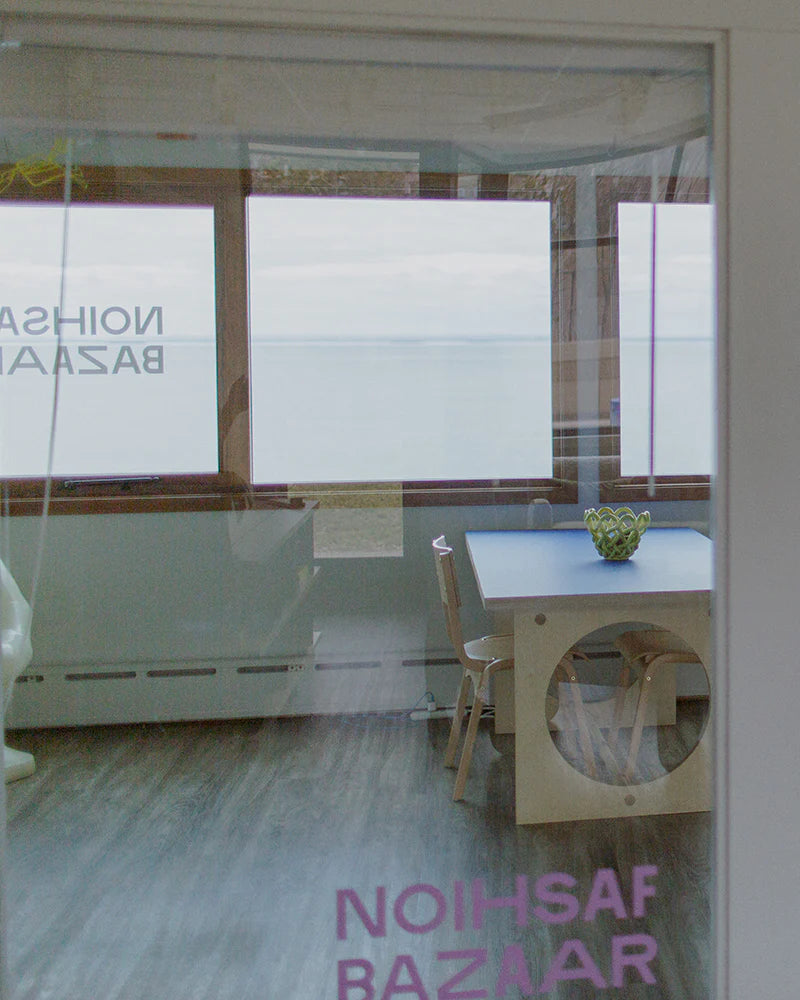 View through glass door showing a room with a table and chairs, a bowl on the table, and large windows overlooking a body of water. Signs with the text NOIHSAF BAZAAR are on the door.