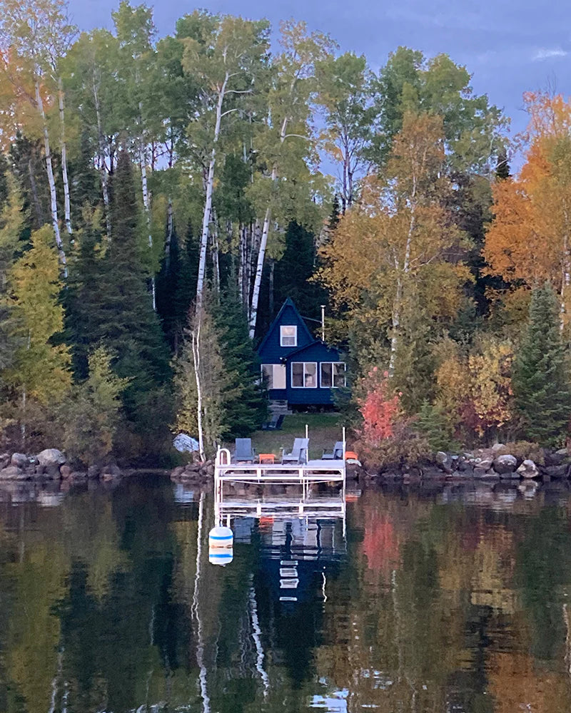 A small cabin with a porch is nestled among trees with autumn foliage, facing a calm lake with a dock and buoy in the foreground.