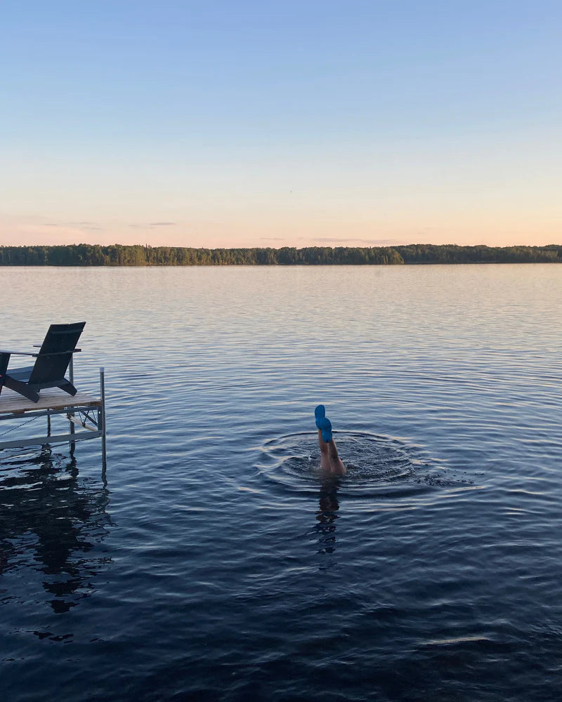 Person in a blue swim cap swimming in a lake near a small dock with two loungers, under a clear sky.