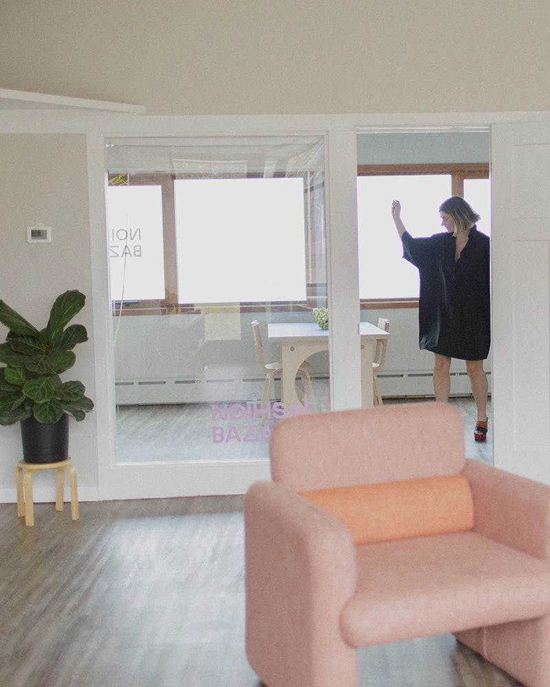 Woman in a black dress stands in a room with large windows, near a table and chairs. A pink chair and a potted plant are in the foreground.