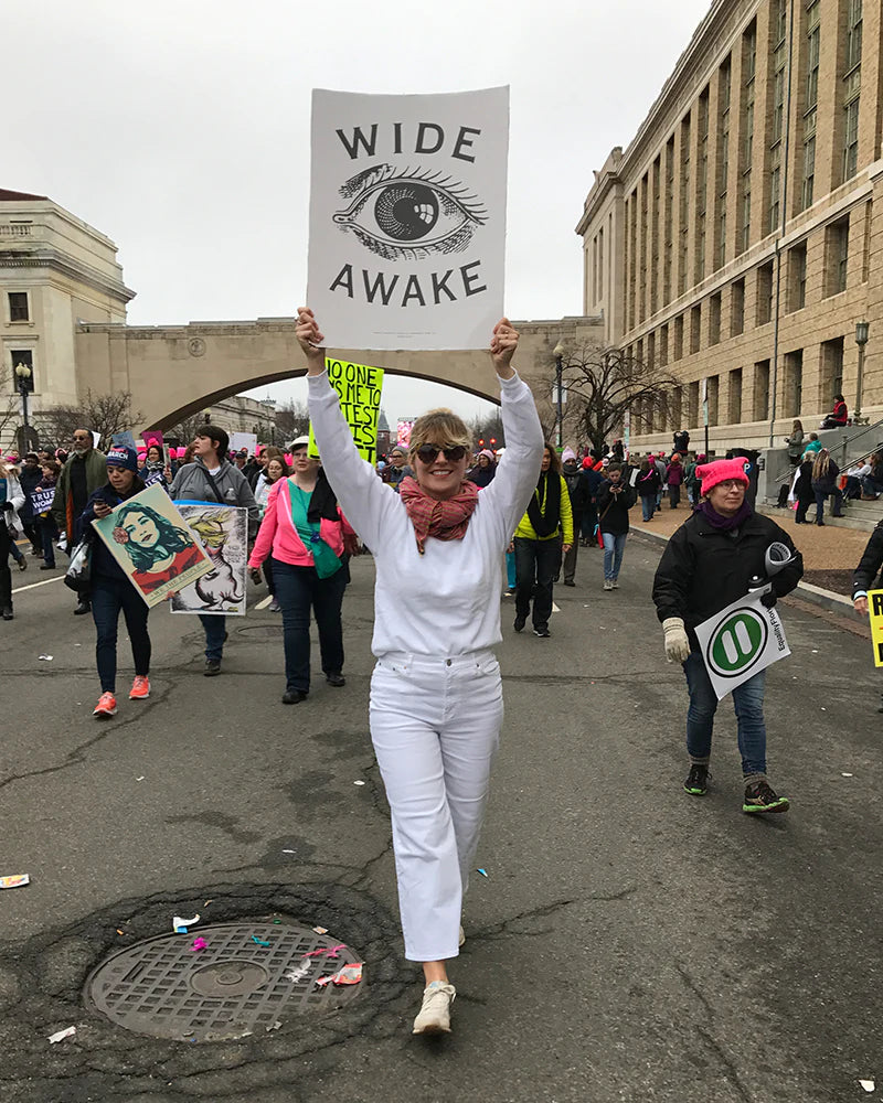 Person in white outfit holding a Wide Awake sign at a protest. Others are visible holding signs and walking on the street.