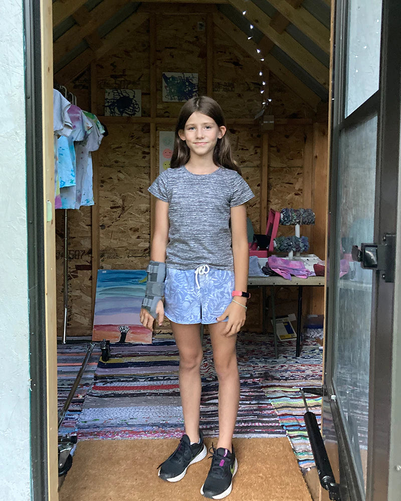 A girl wearing a gray shirt and blue shorts stands inside a wooden room with colorful fabrics and decorations. She has a brace on her left arm and is wearing black sneakers.