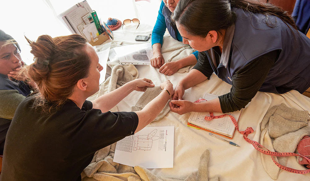 People sitting around a table, examining fabric and knitting patterns. Tools like a measuring tape, notebook, and papers are visible.