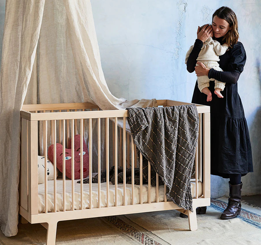 Woman in a black dress holds a baby next to a wooden crib with a draped cloth and stuffed toys inside, set against a textured wall.