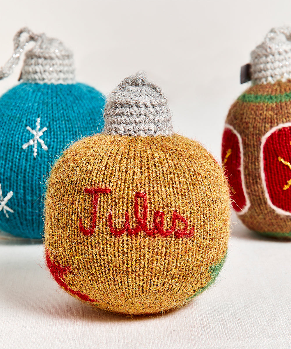 A personalized gold ornament with "Jules" embroidered, displayed in front of blue and brown knitted Christmas ornaments.