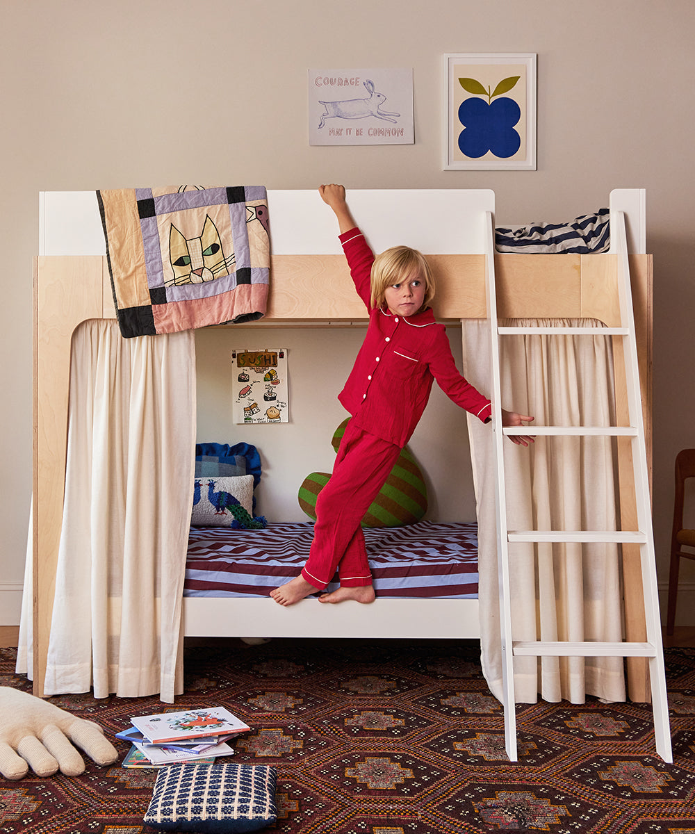 A child in red pajamas hangs from a Perch Bunk Bed with Curtains in a cozy, colorful bedroom filled with toys and books.
