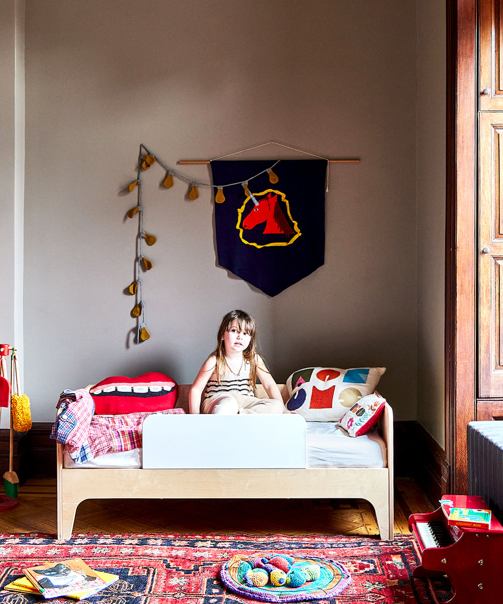 A girl sits on a bed in a colorful bedroom decorated with toys and a Light Bulb Garland hanging on the wall above her.
