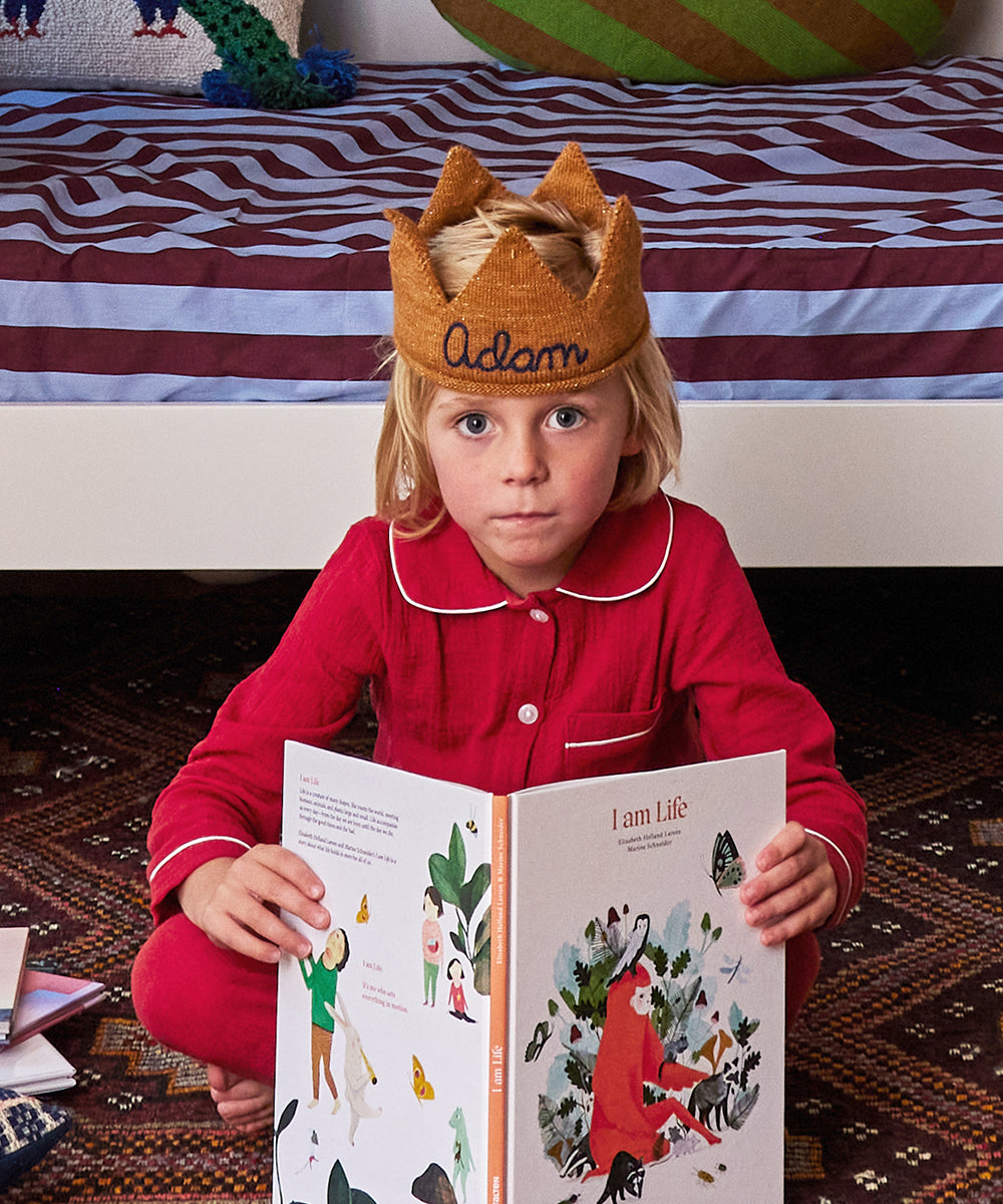 A child in red pajamas, wearing a Personalized Gold Crown - Indigo, reads "I am Life" on a patterned rug beside a bed.