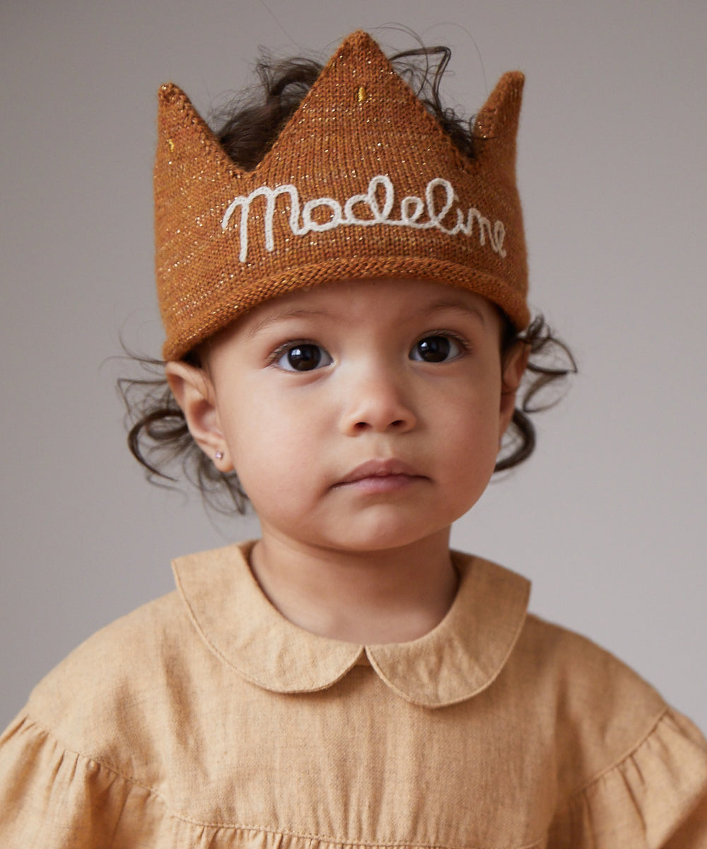 Toddler with curly hair wears a Personalized Gold Crown labeled “Madeleine” and a matching dress with Peter Pan collar, facing forward against a plain background.