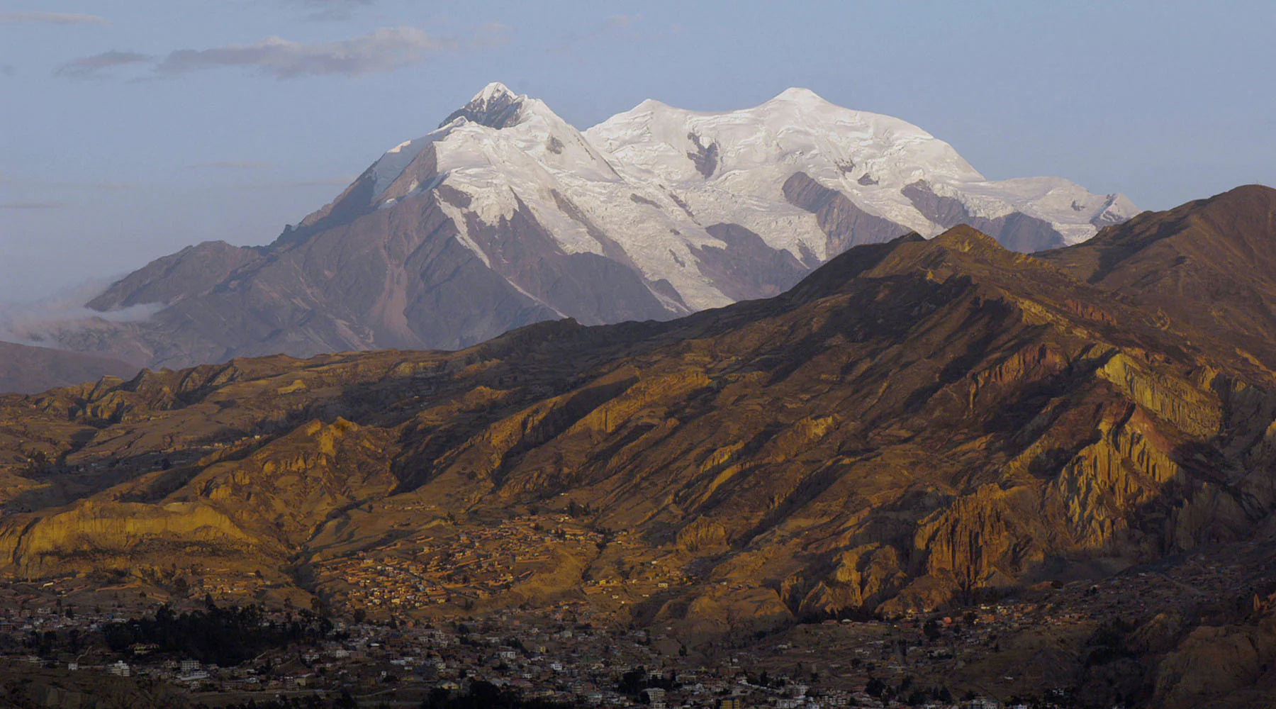 Snow-capped Mount Illimani overlooking a rugged landscape and a small town in the foreground at dusk.