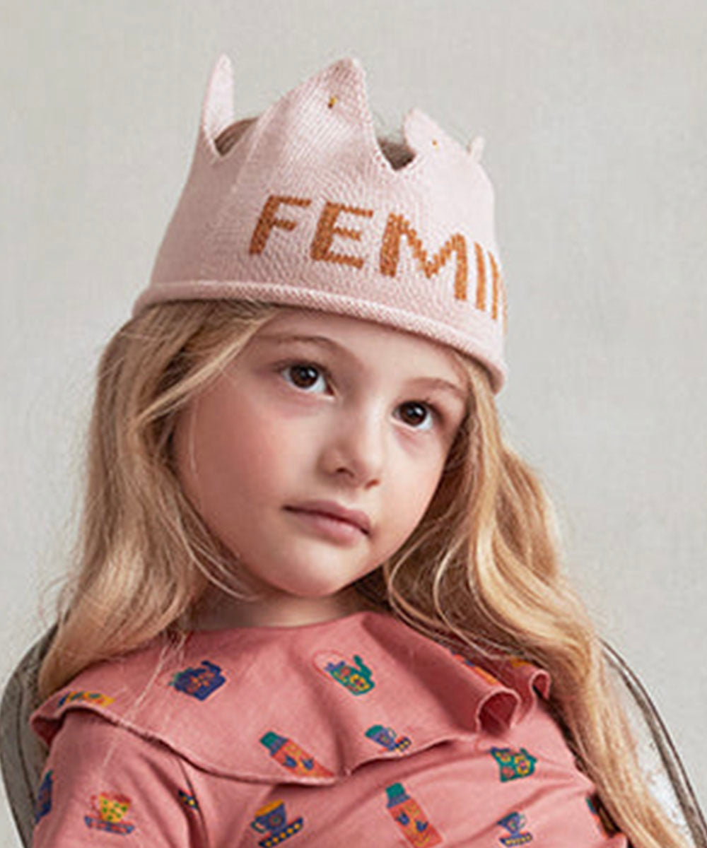 Young girl wearing the Feminist Crown, a pink hat with "FEMIN" on it, paired with a pink dress featuring colorful patterns.
