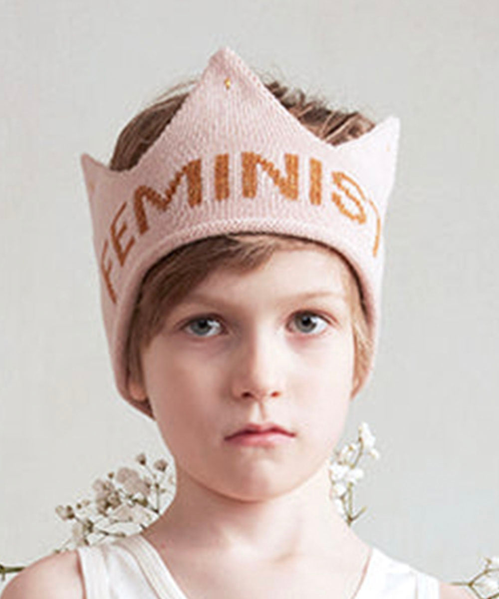 Young child wearing the Feminist Crown—a pink, crown-shaped hat with "FEMINIST"—looks directly at the camera.