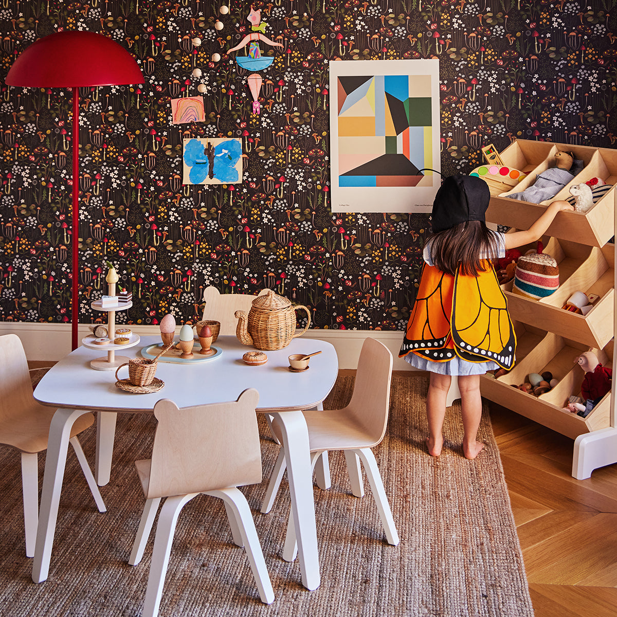 Child in butterfly costume stands near a wooden hexagonal shelf in a playroom with colorful walls, a small table and chairs, toys, and artwork.