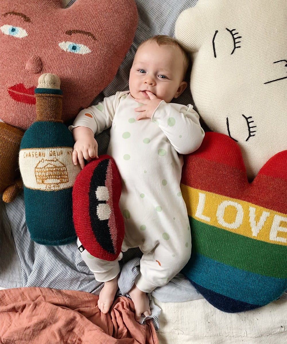 Baby in polka dot onesie lies on bed, surrounded by colorful face-shaped pillows and a Heart Pillow - Rainbow.