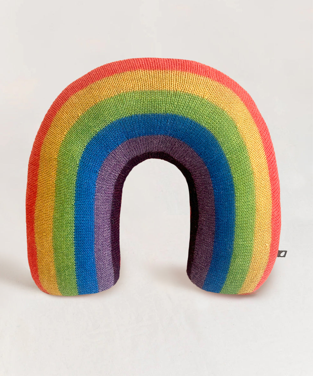 The Rainbow Pillow is a rainbow-shaped cushion with colorful stripes, set against a plain light background.