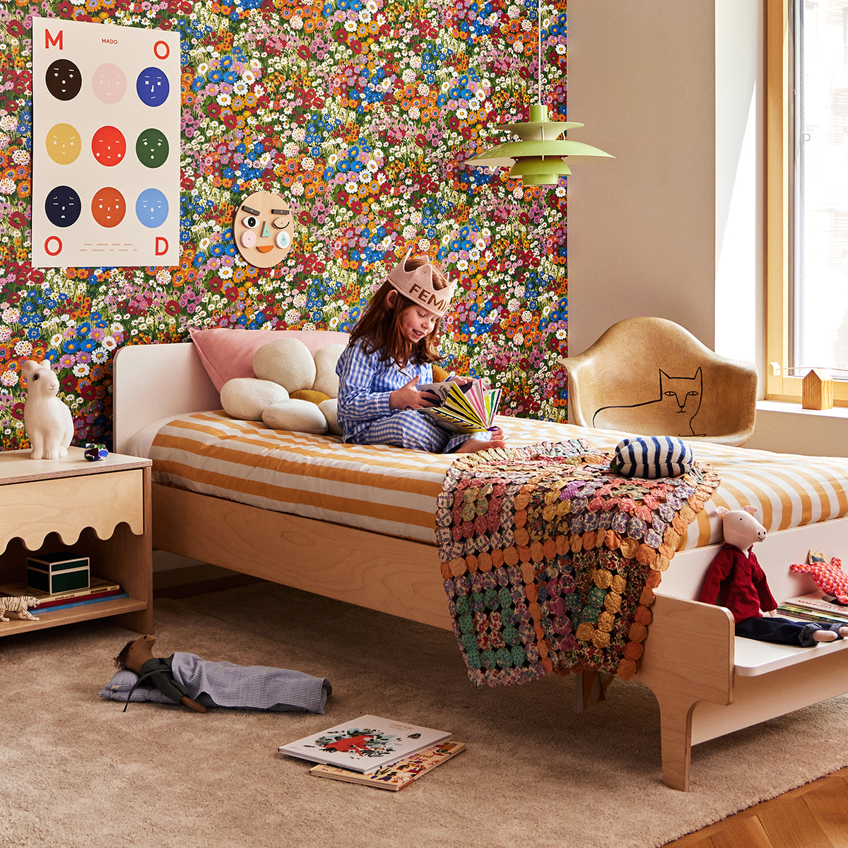 A child sits on a striped bed in a colorful room, drawing with markers. The room features floral wallpaper, a wooden nightstand, and a chair with a cat design.