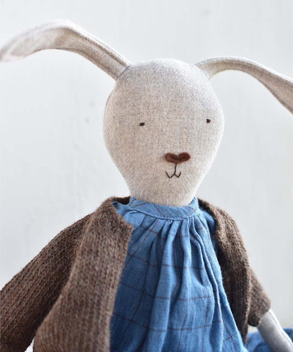 Ruthie le Rabbit: a beige stuffed bunny with long ears, simple stitched face, blue dress, and brown cardigan, set against a plain light background.