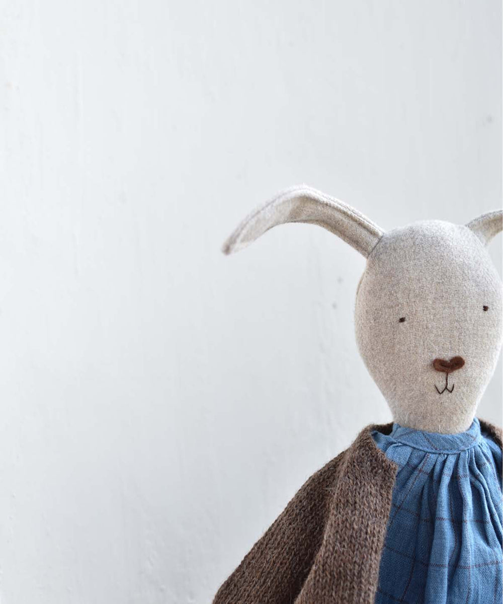 Ruthie le Rabbit plush doll in a blue dress and brown sweater, posed against a plain light gray background.