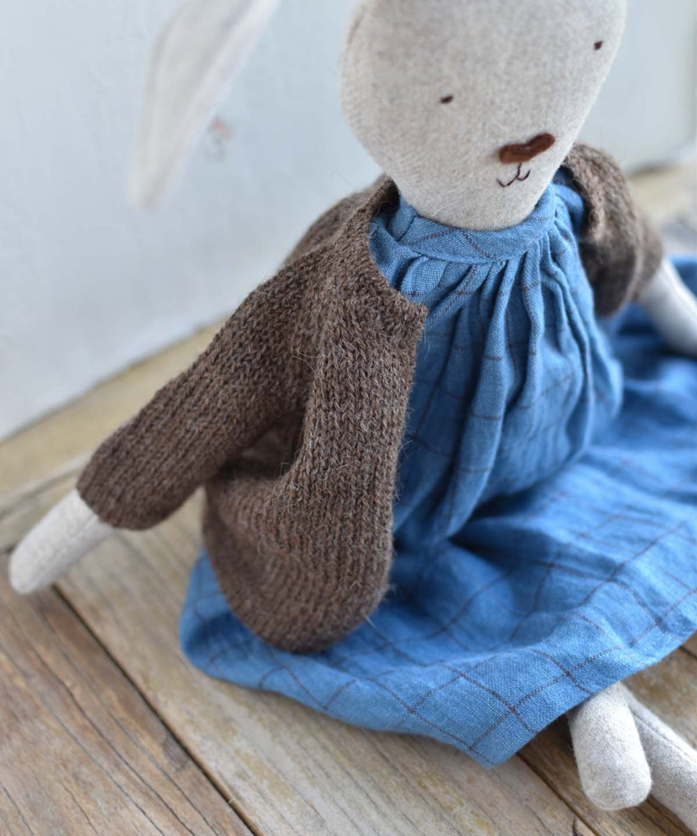 Ruthie le Rabbit, in a blue dress and brown knit cardigan, sits on a wooden surface by a white wall.