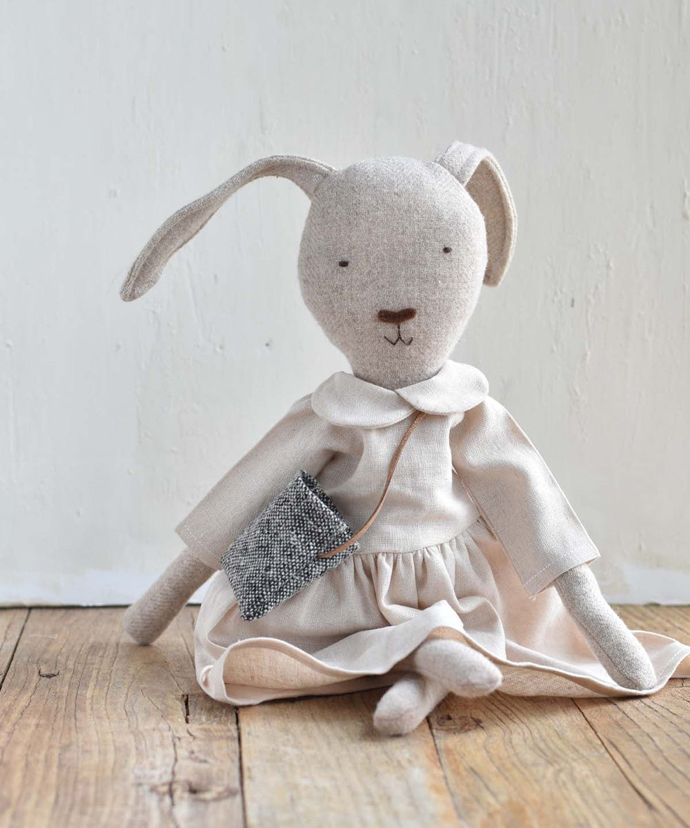 Ruthie le Rabbit – Linen Dress: Stuffed bunny doll in a linen dress with a small gray purse, sitting on a wooden floor.