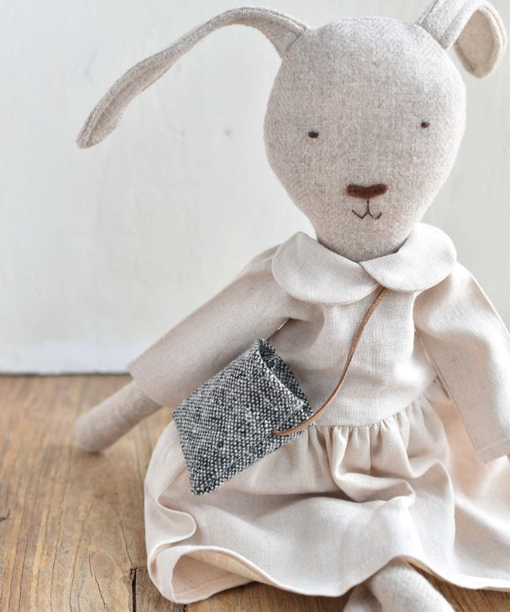 Ruthie le Rabbit – Linen Dress sits on a wooden floor in a cream dress with a gray bag, against a white wall.