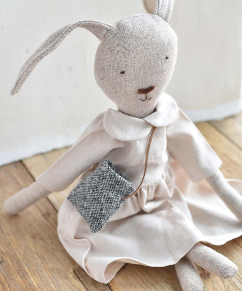 Ruthie le Rabbit – Linen Dress: Plush rabbit doll in a linen dress with a gray bag, sitting on a wooden floor.