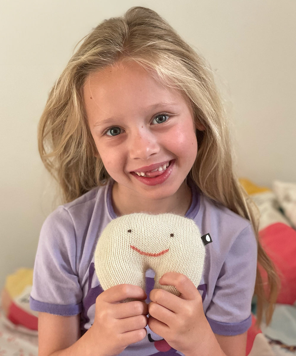 A smiling girl with long blonde hair holds a Tooth Fairy Pillow and shows the gap from her missing tooth.