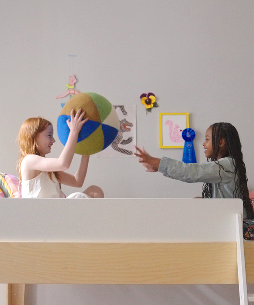 Two girls sit on a bed in a bright, decorated room, playing catch with the Circus Ball - Royal Blue.