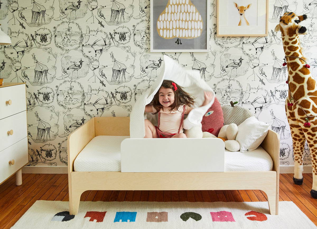 Child happily playing on a bed in a decorated room with animal-themed wallpaper, a large toy giraffe, and a colorful rug.