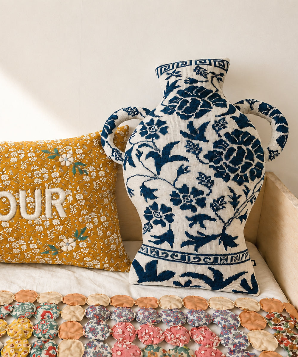 The Porcelain Vase Pillow, featuring blue and white floral patterns, sits beside a yellow floral pillow on a quilted surface.