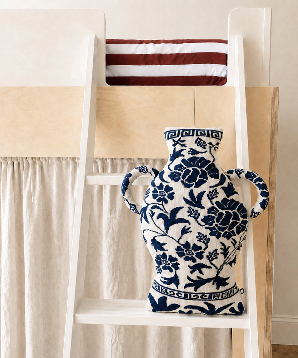 The Porcelain Vase Pillow in blue and white embroidery rests on a white ladder before a bunk bed with red-and-white striped bedding.