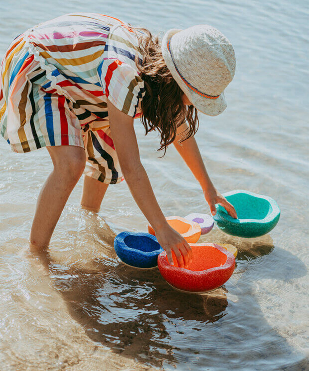 A child in a striped dress and hat plays with the Le Inside Set, colorful bowls, in shallow beach water.