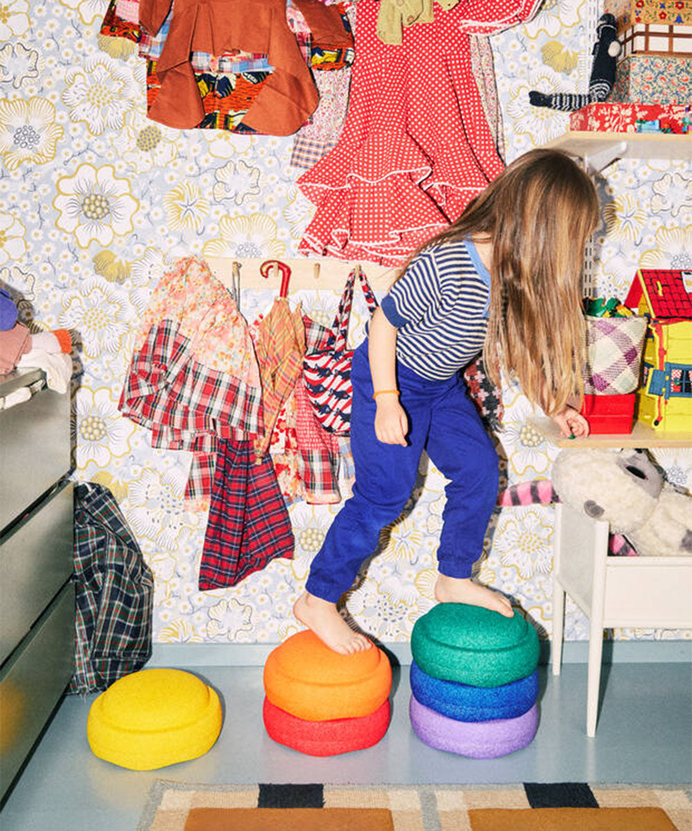 A child steps on the Le Original Rainbow colorful cushions in a playroom with bright clothes and patterned wallpaper.