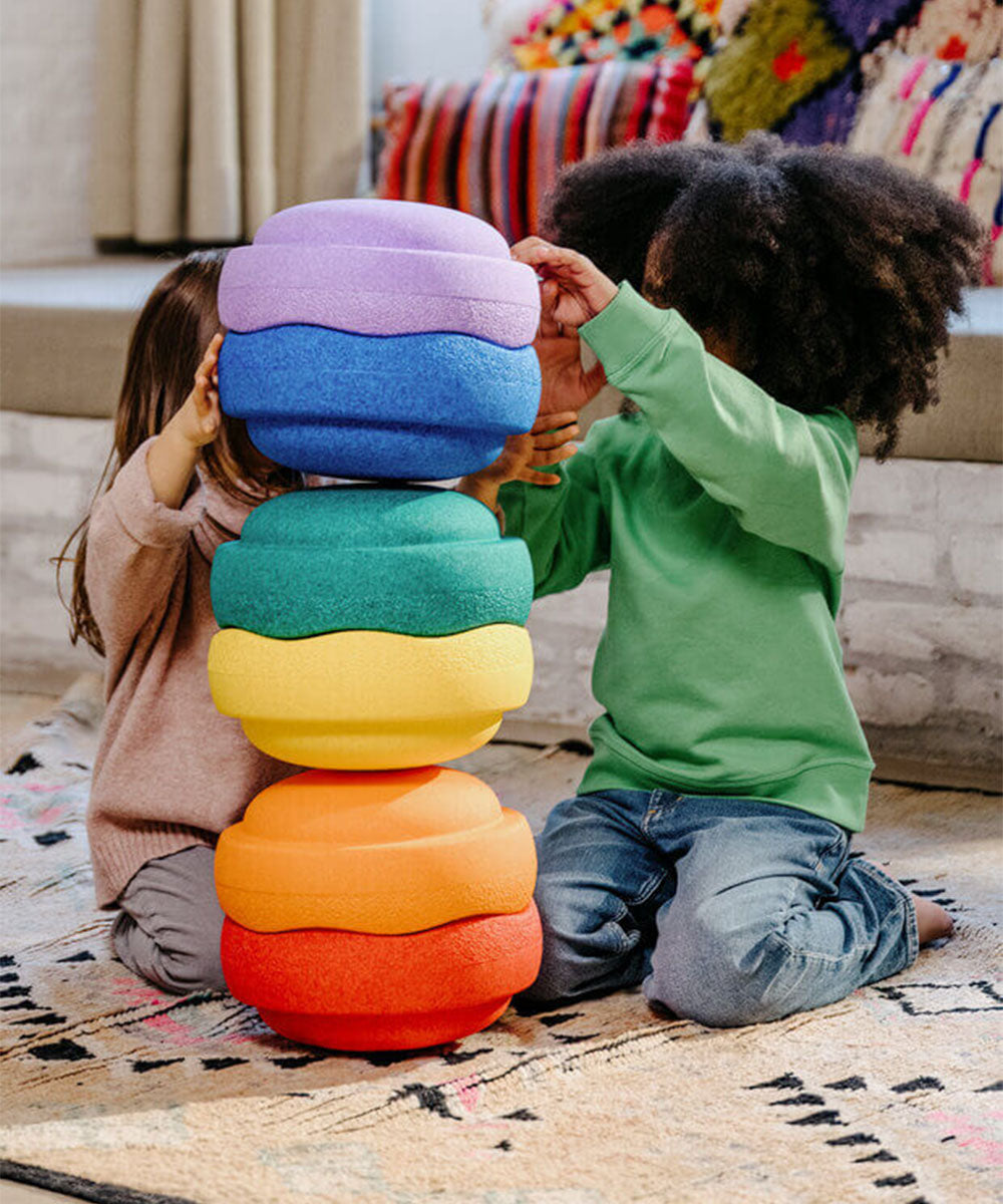 Two children stack Le Original Rainbow foam rings while playing on a patterned rug indoors.